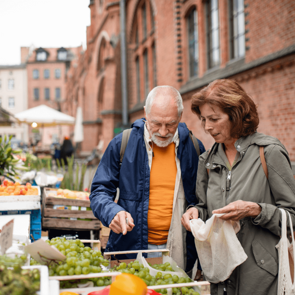 An elderly couple shops for fresh produce at an outdoor market, examining grapes displayed at a vendor's stall. - Home Instead