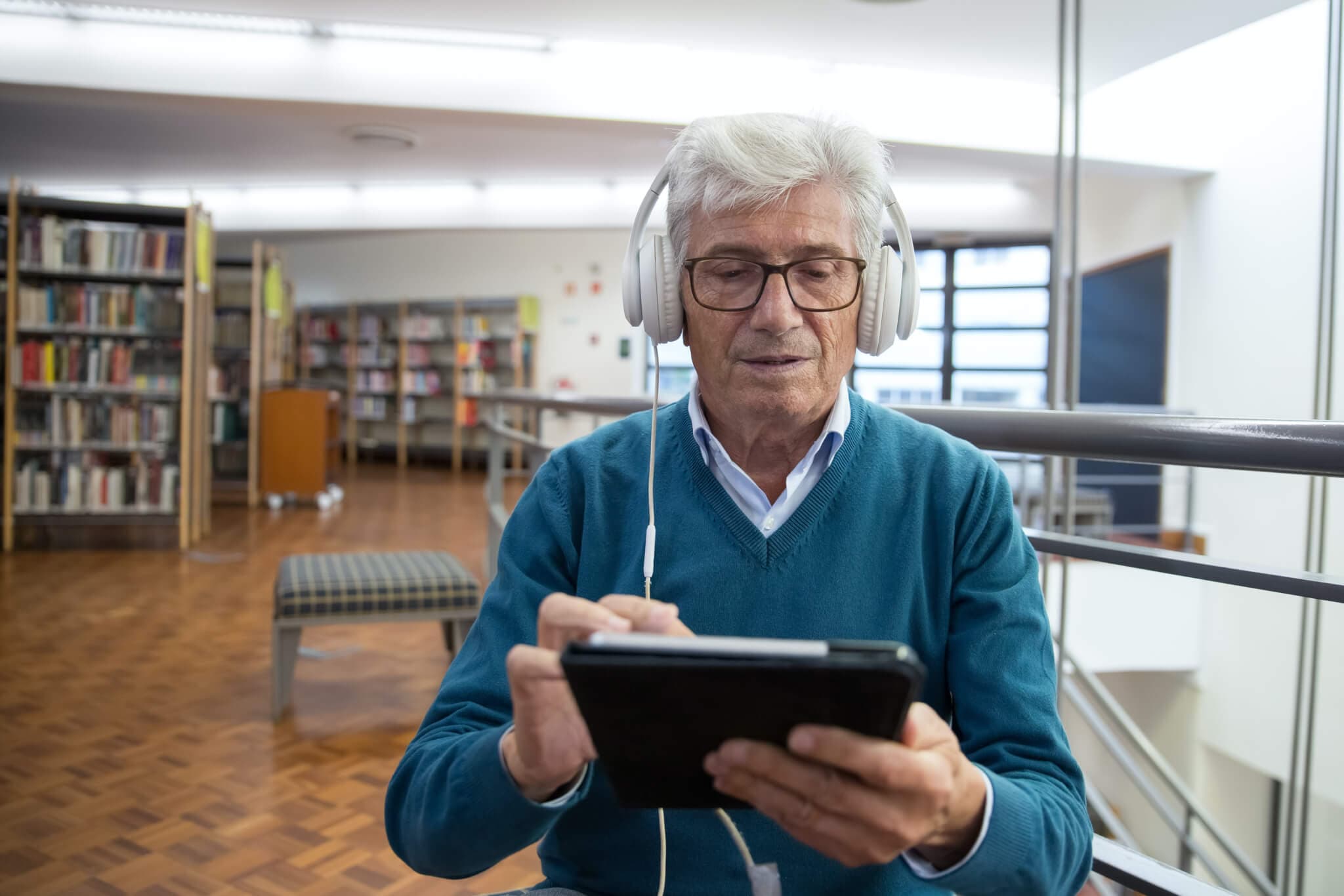 Older man in a library using a tablet while wearing headphones. Bookshelves and a bench are visible in the background. - Home Instead