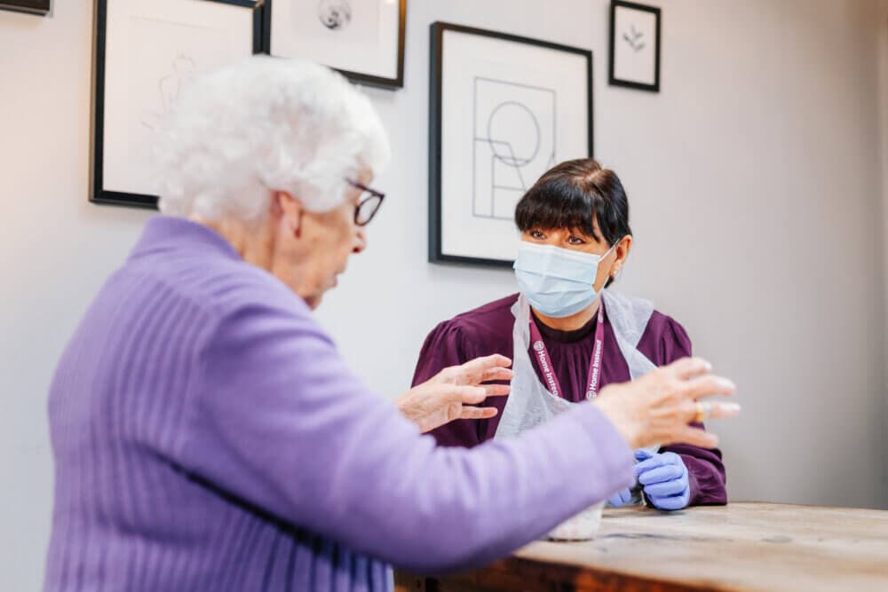 Elderly woman and masked caregiver talking at a table in front of framed wall art. - Home Instead