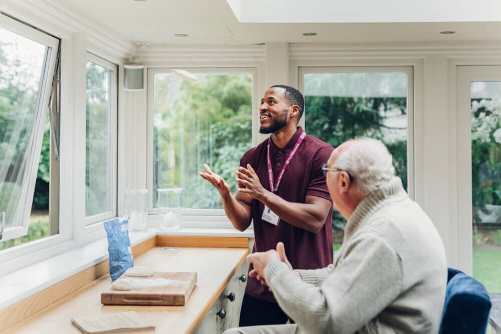 A man in a polo shirt talks to an elderly man at a kitchen counter with a cutting board and ingredients. - Home Instead