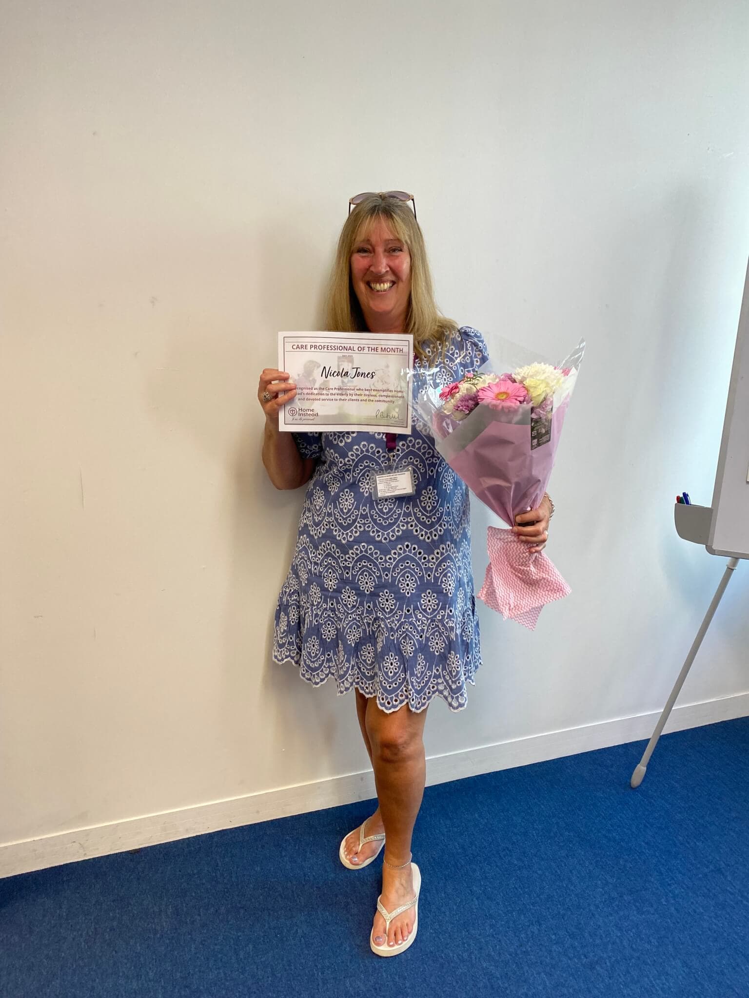 A person in a blue dress holds a certificate in one hand and a bouquet of flowers in the other, smiling. - Home Instead