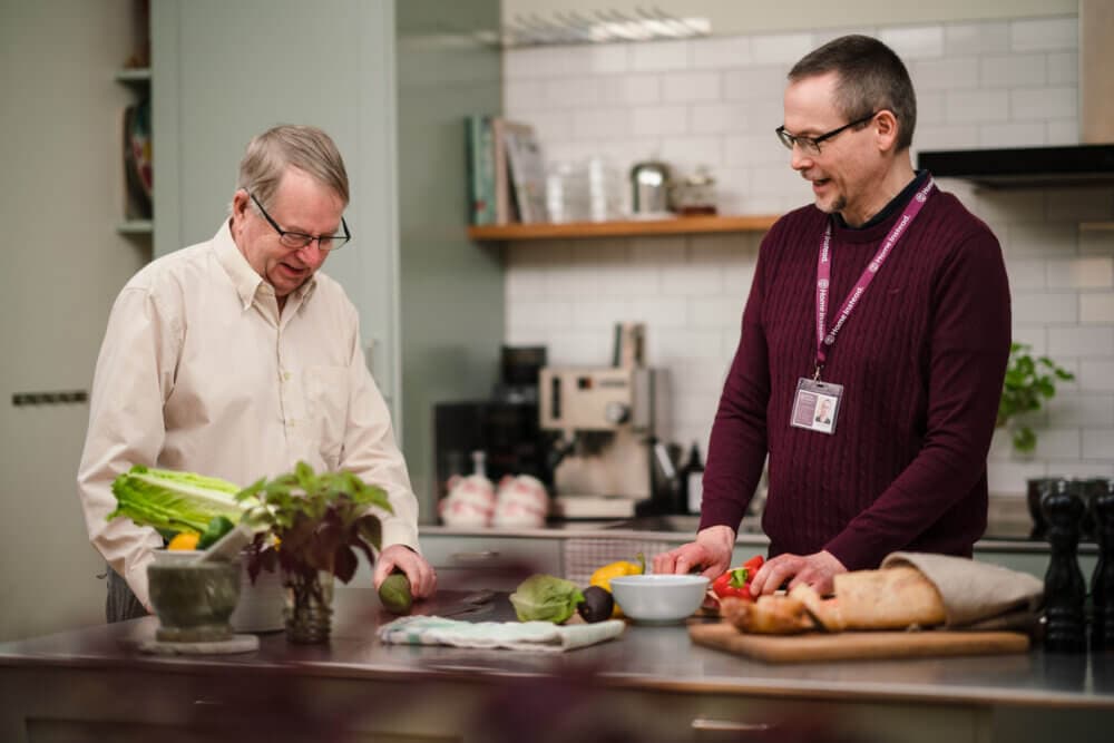 Two men are preparing food together in a modern kitchen, surrounded by fresh vegetables and bread on the counter. - Home Instead