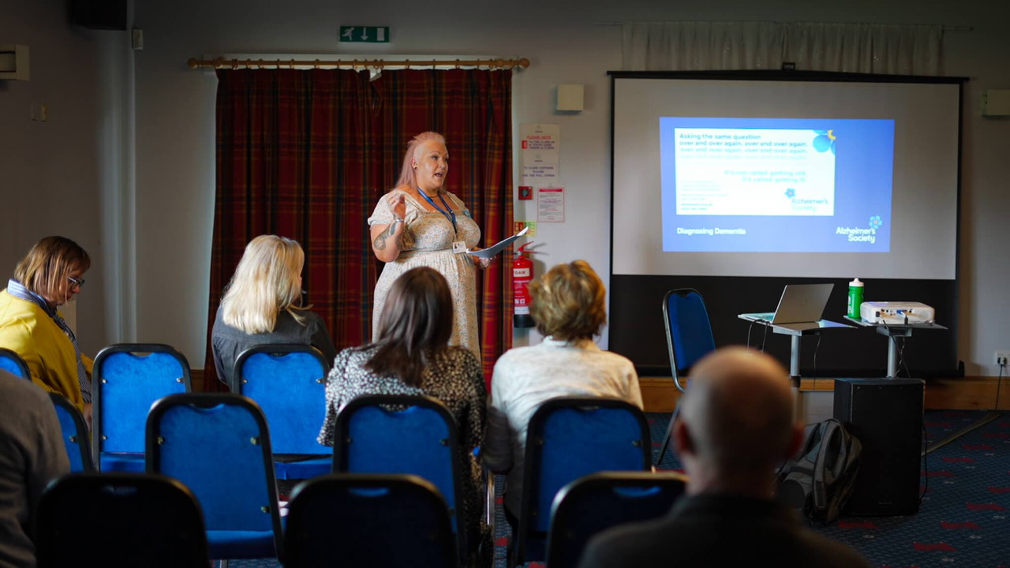 A woman is giving a presentation to a small audience in a seminar room with a screen displaying information. - Home Instead