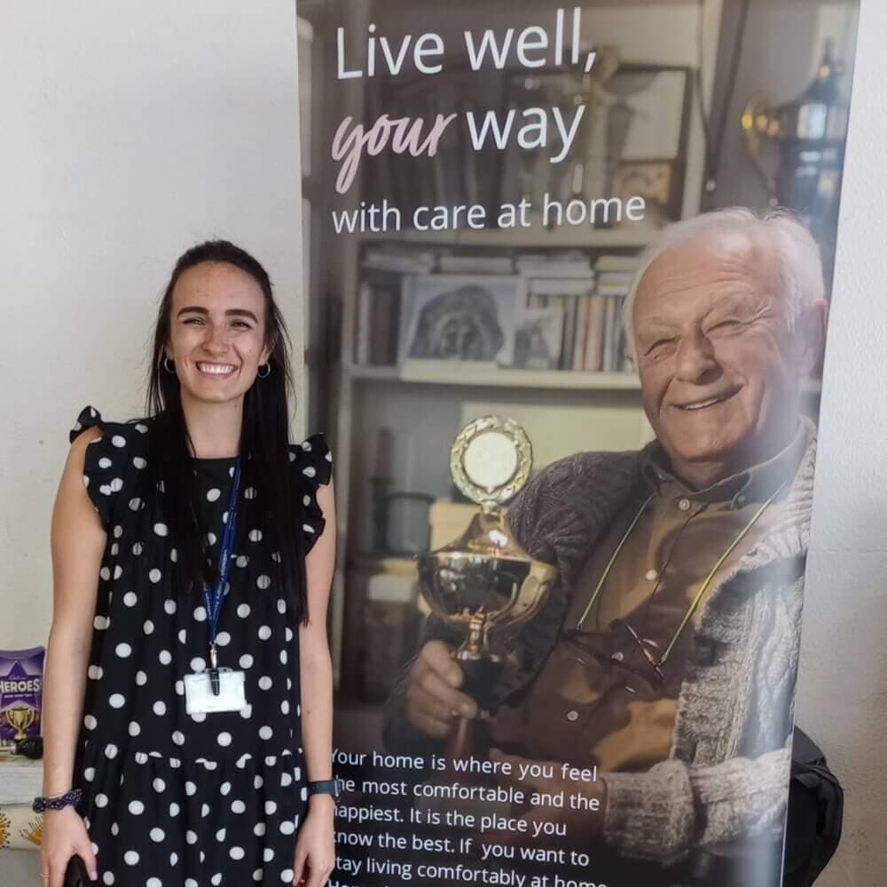 A woman in a polka dot dress stands beside a banner showing an elderly man holding a trophy. - Home Instead