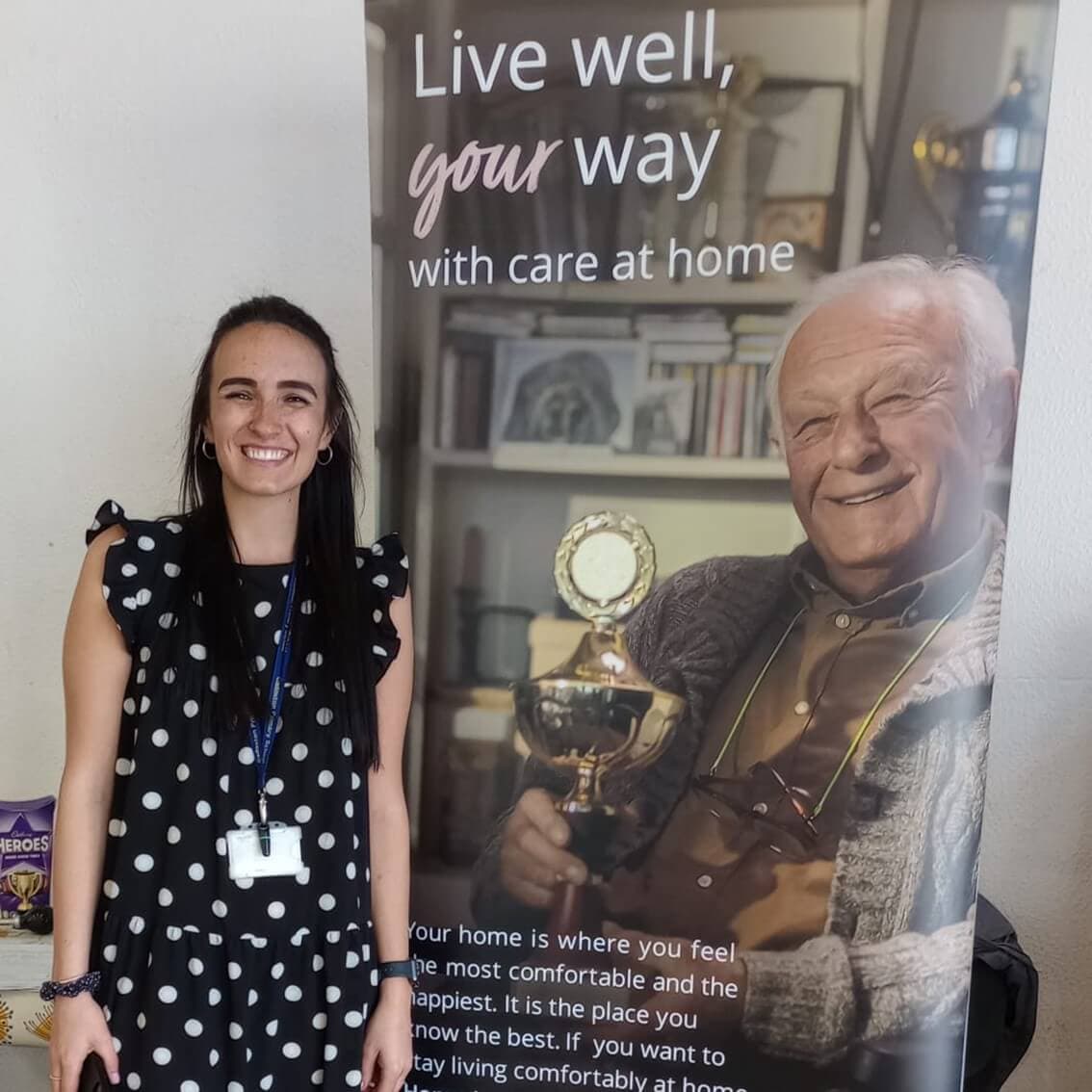 A woman in a polka dot dress stands beside a banner showing an elderly man holding a trophy. - Home Instead