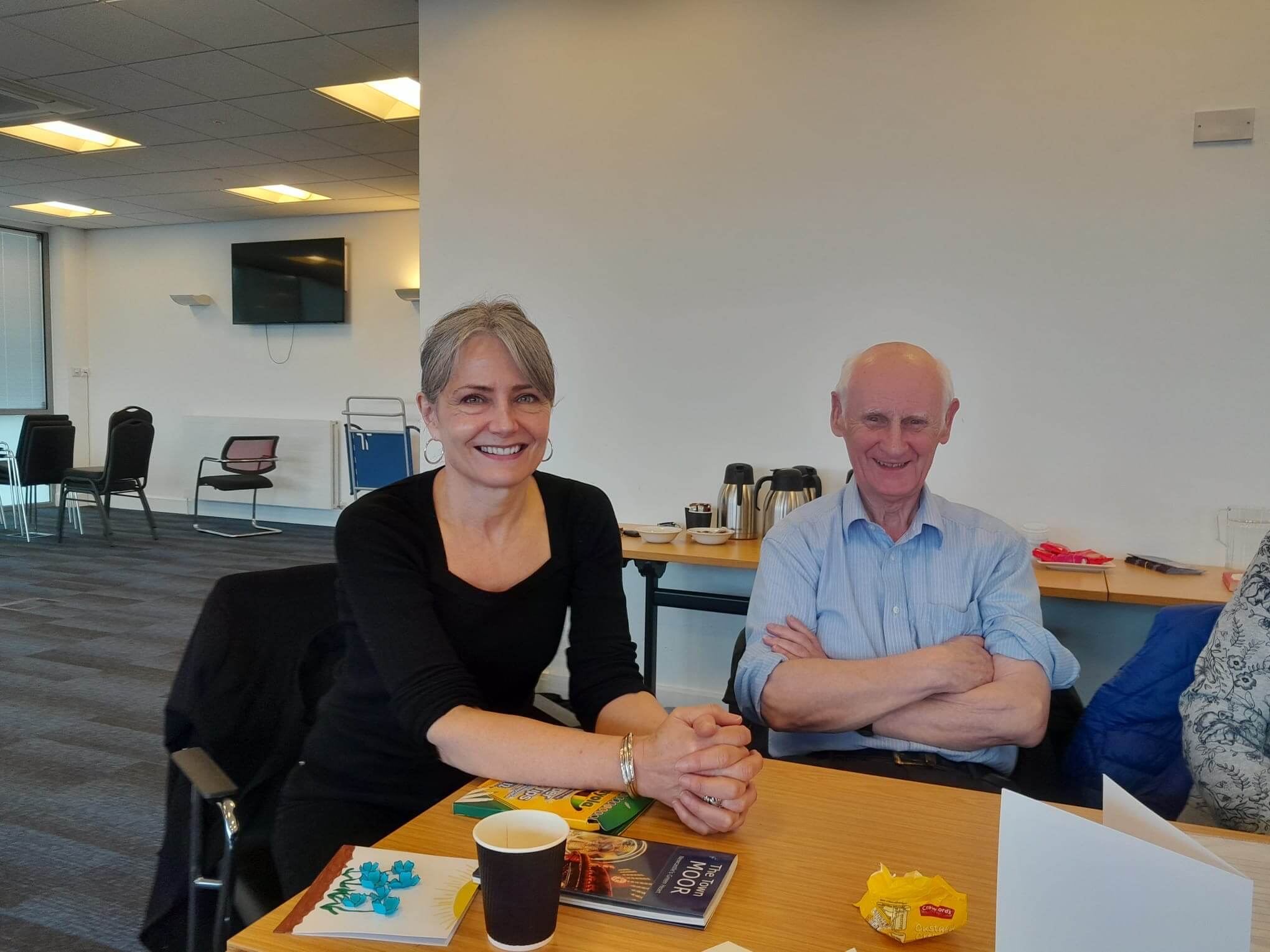 Two people seated at a table, smiling, with coffee cups and books in front of them in a brightly lit room. - Home Instead