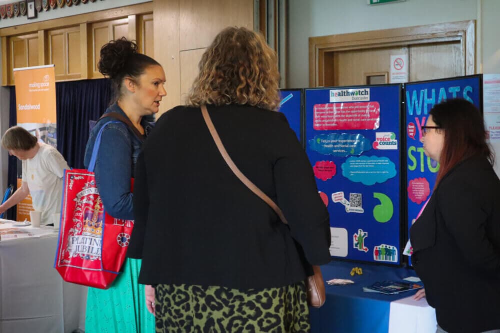 Three women engage in conversation at an information booth during an indoor event. - Home Instead