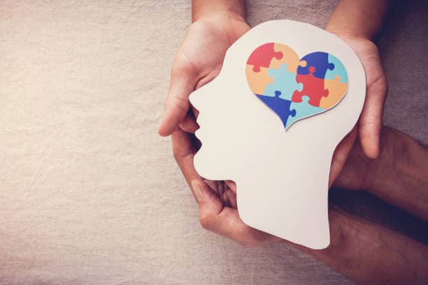 Hands holding a cutout of a head with a colorful heart-shaped puzzle inside, symbolizing mental health and autism awareness. - Home Instead