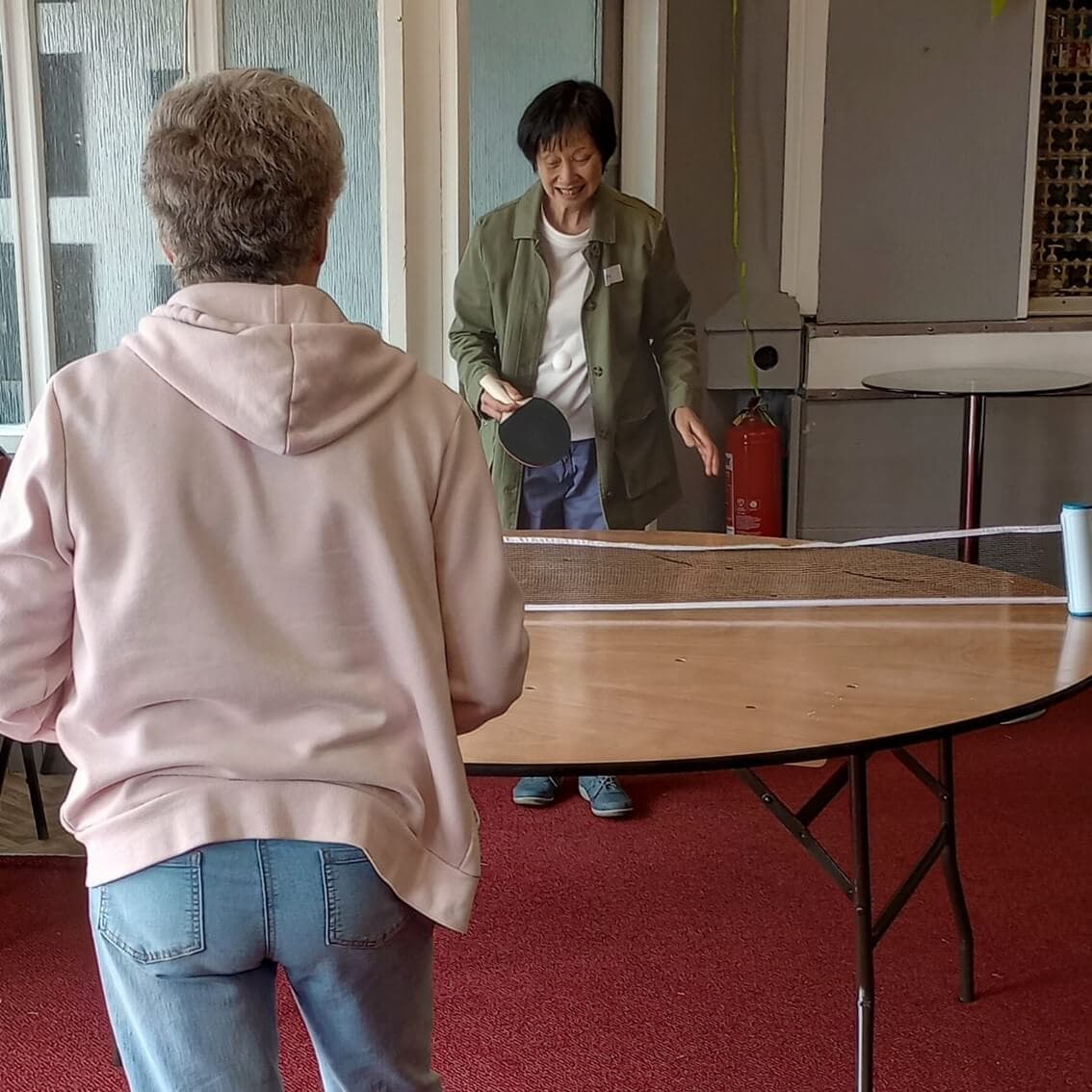 Two women playing table tennis indoors using a makeshift setup on a wooden table. One is seen from the back. - Home Instead