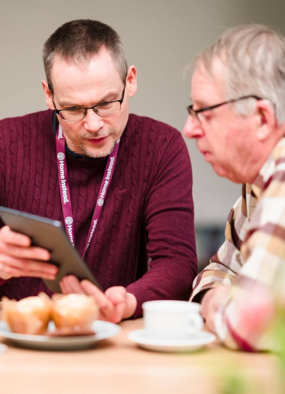 A Care Professional helps a client with a tablet at a table with muffins and a coffee cup. - Home Instead Bournemouth & Christchurch