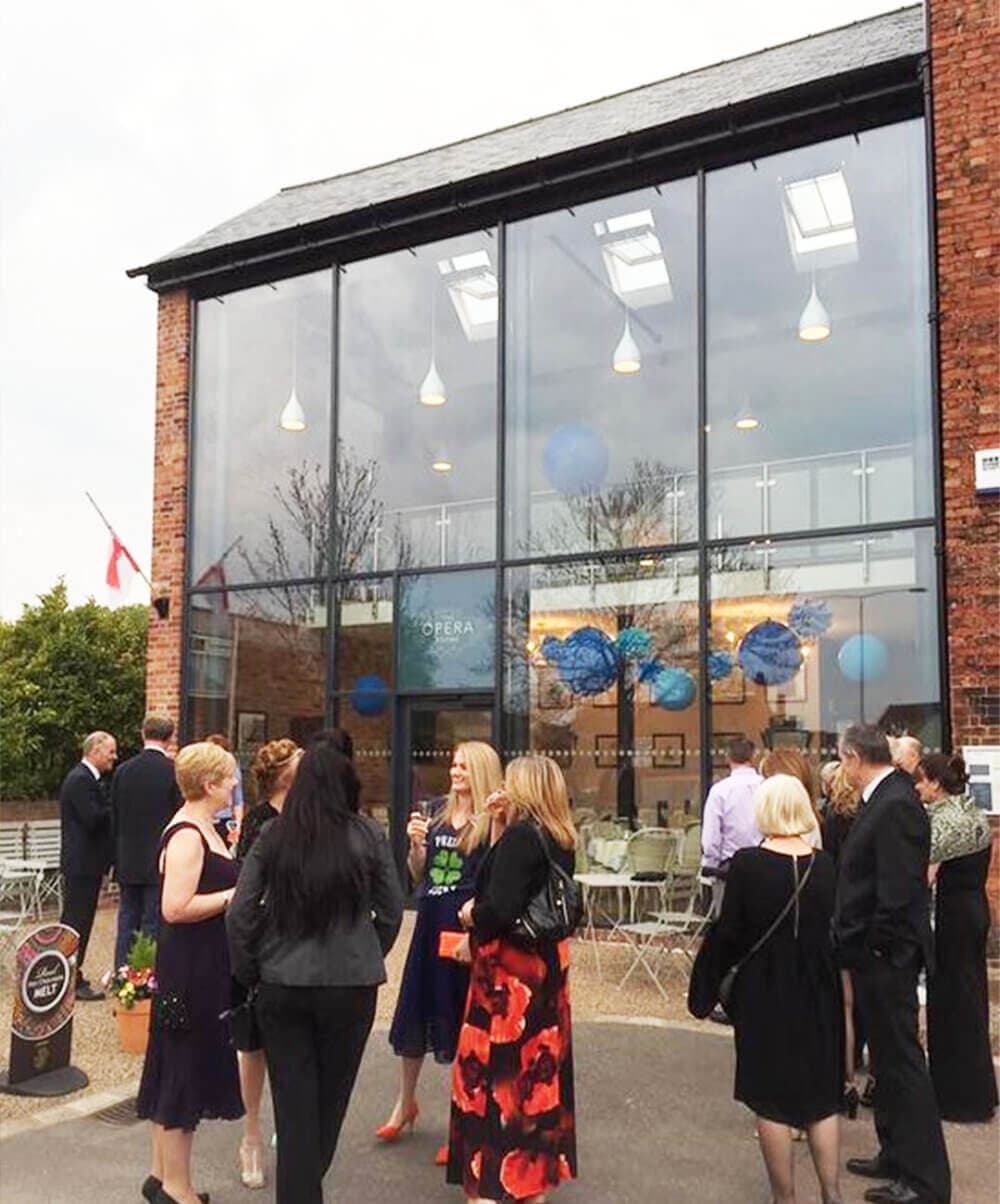 A group of people in formal attire socialize outside a two-story brick building with large windows. - Home Instead