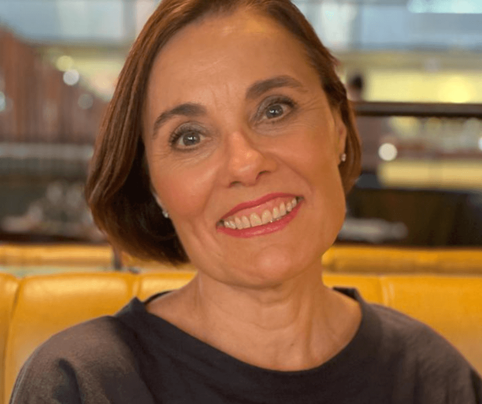 A woman with short brown hair and a gray sweater smiles while sitting on a yellow sofa in a restaurant. - Home Instead