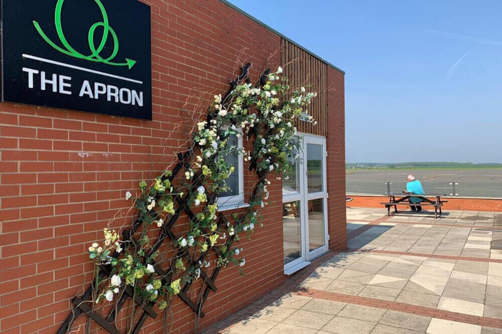 A café with a flower trellis and seating outside overlooks an empty tarmac under a clear sky. - Home Instead
