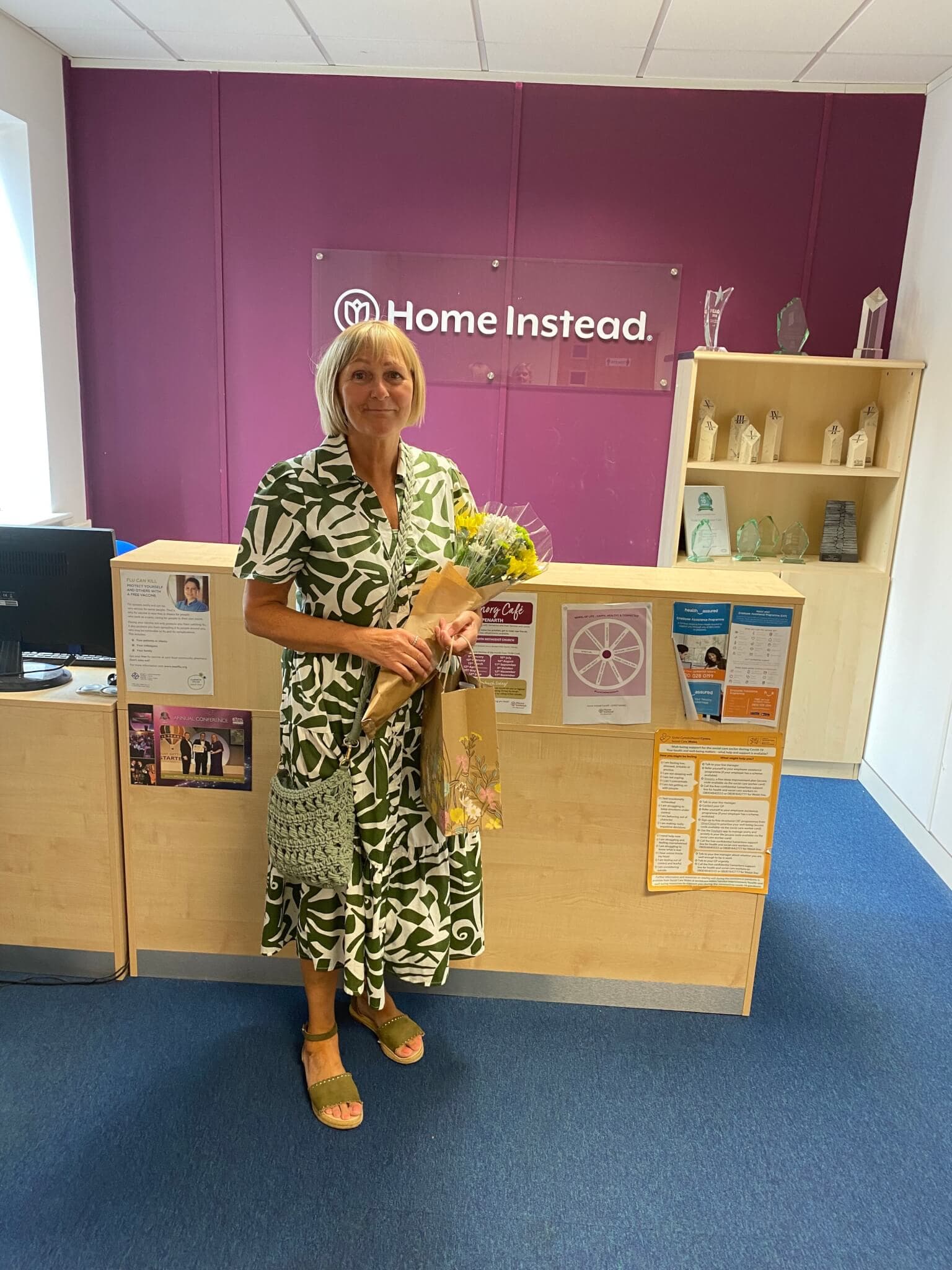 A woman in a green dress stands in front of a desk at an office, holding flowers and a gift, with a "Home Instead" sign behind her. - Home Instead