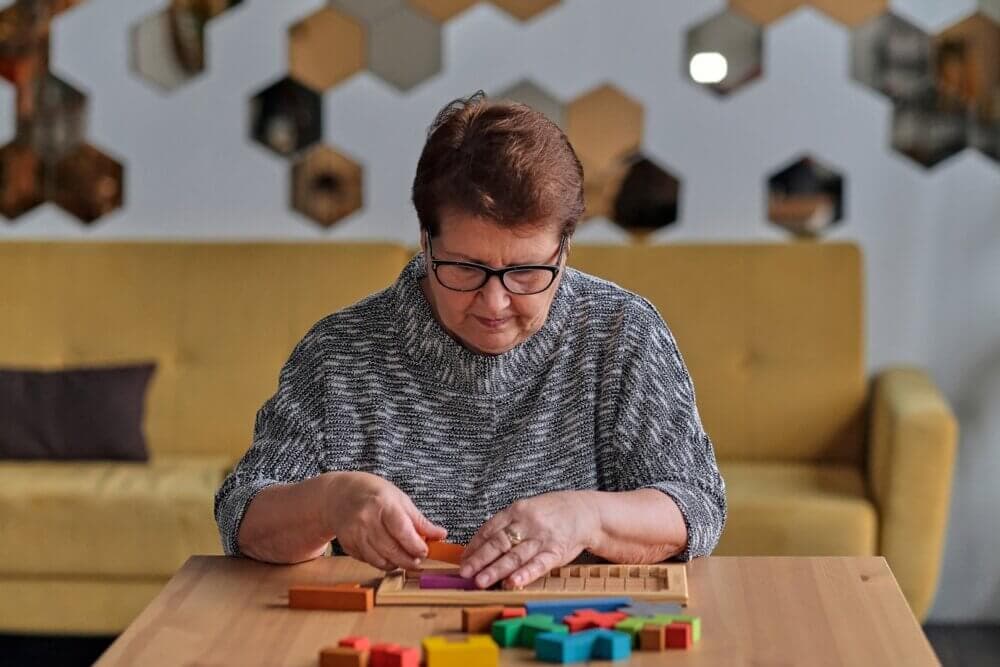 Older woman with glasses concentrating on building a structure with colorful wooden blocks at a table. - Home Instead