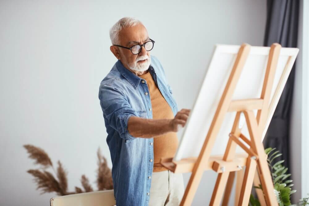 Elderly man with glasses and gray hair painting on a canvas in a well-lit room with plants in the background. - Home Instead