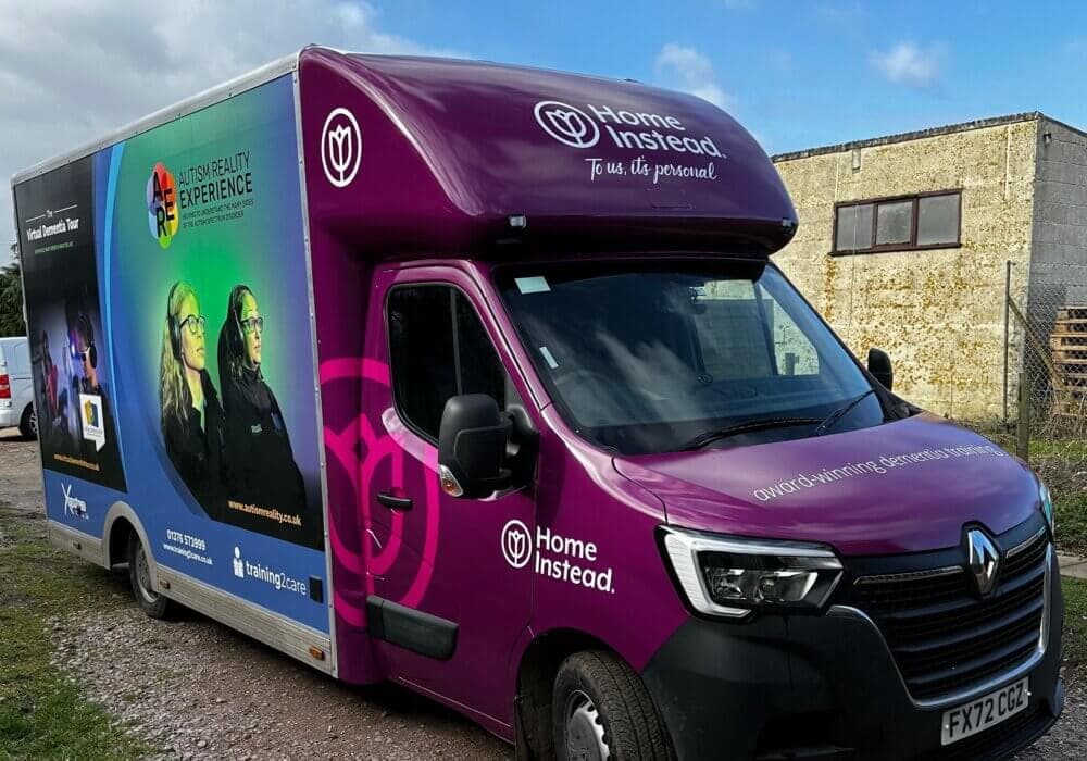 A purple and blue truck with "Home Instead" branding and "Autism Reality Experience" graphics. License plate FX72 CCJ. - Home Instead