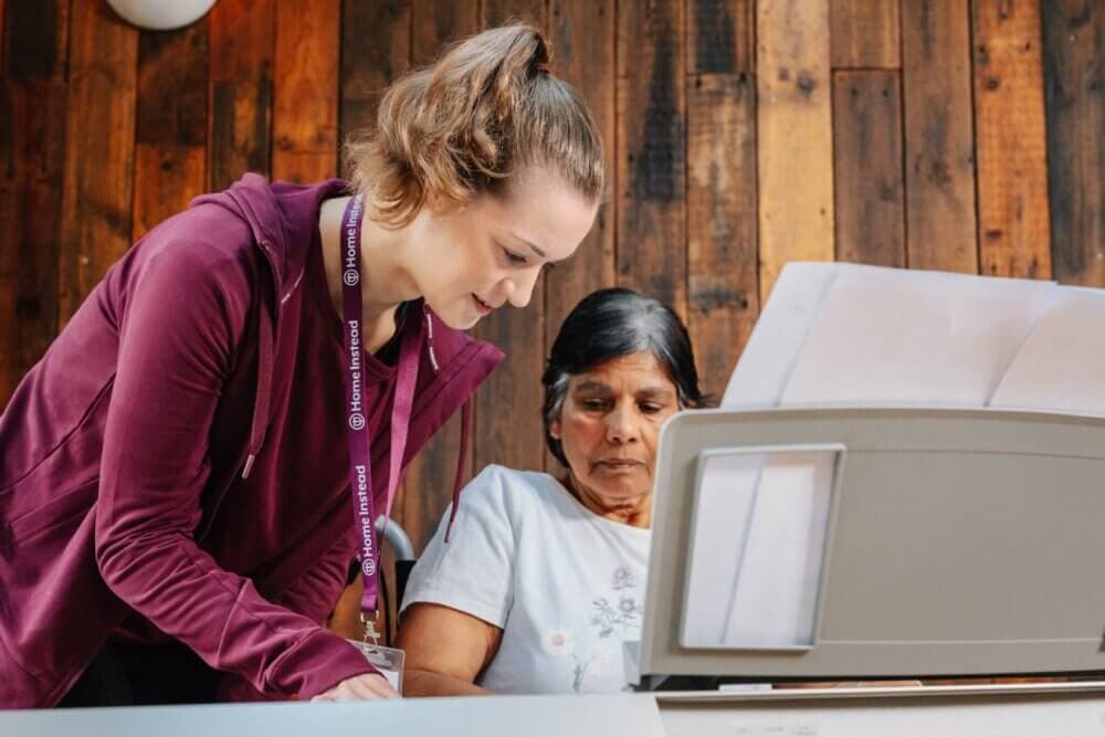 A young woman assists an older woman with paperwork at a table inside a wooden-paneled room. - Home Instead