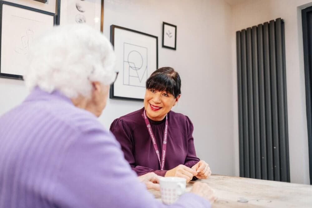 A woman with dark hair speaks to an elderly woman with white hair at a table in a modern room. - Home Instead