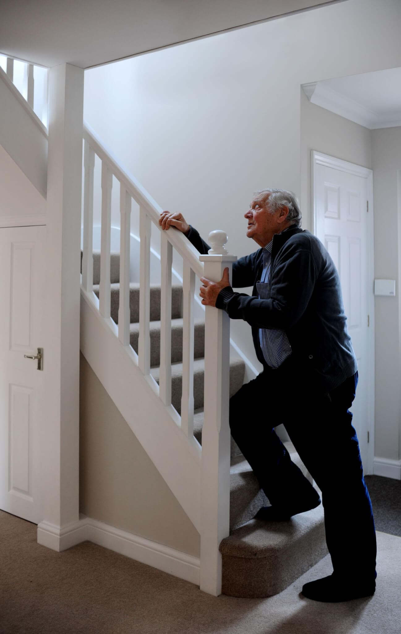 An elderly man holds onto a stair railing while climbing carpeted stairs inside a well-lit home. - Home Instead