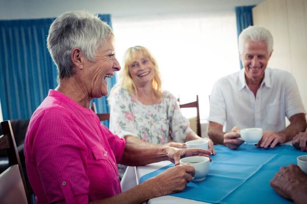 Three elderly people smiling and enjoying coffee around a table with a blue tablecloth in a bright room. - Home Instead