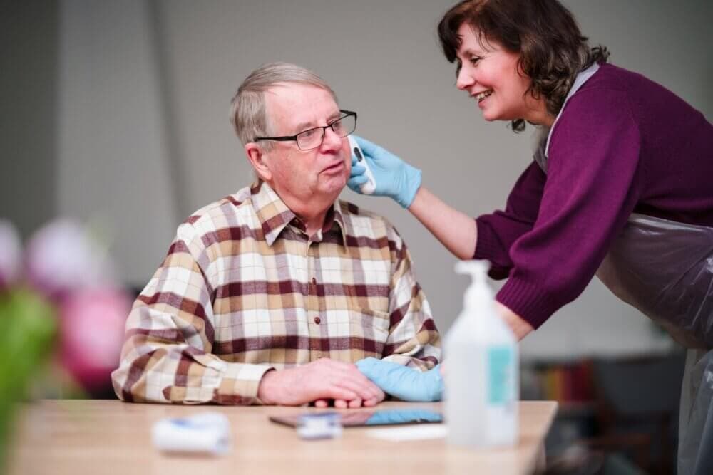 A caregiver in gloves and an apron smiles while assisting an elderly man sitting at a table with a plaid shirt. - Home Instead
