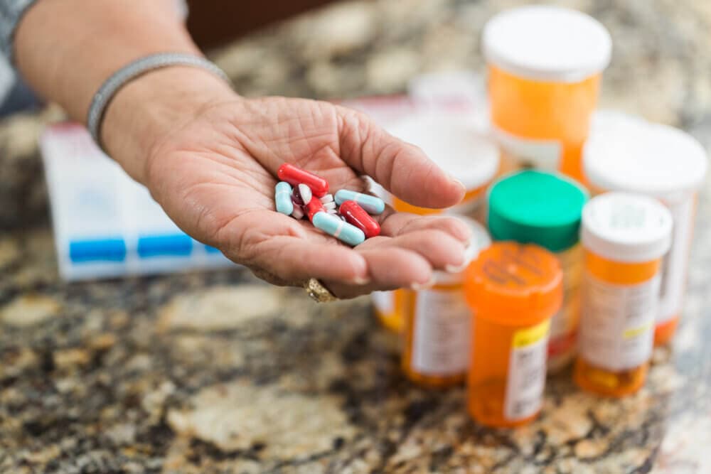 A hand holding various pills with several medication bottles in the background on a countertop. - Home Instead