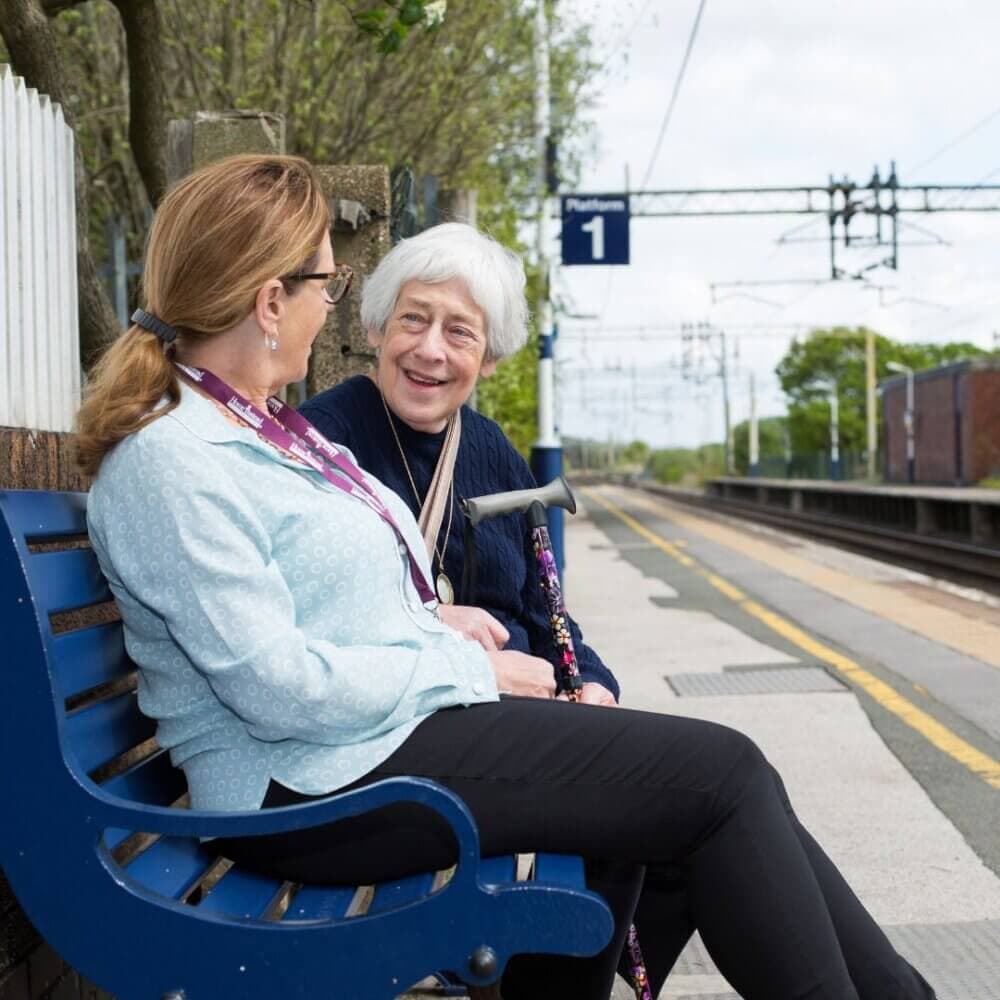 Two women sit on a bench at a railway station, smiling and talking, with train tracks and a Platform 1 sign visible. - Home Instead