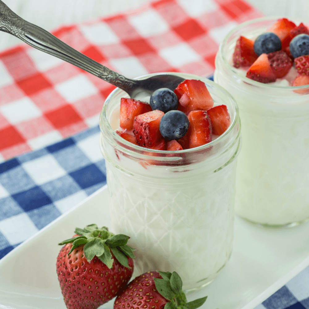 Two jars of yogurt topped with blueberries and strawberries on a checkered cloth, with fresh strawberries beside them. - Home Instead