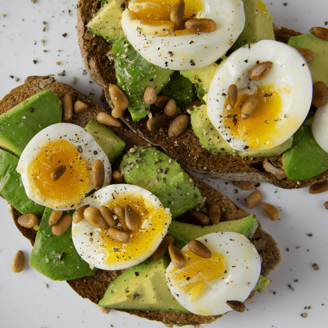 Two toast slices topped with avocado, soft-boiled eggs, pine nuts, and black pepper on a white plate. - Home Instead