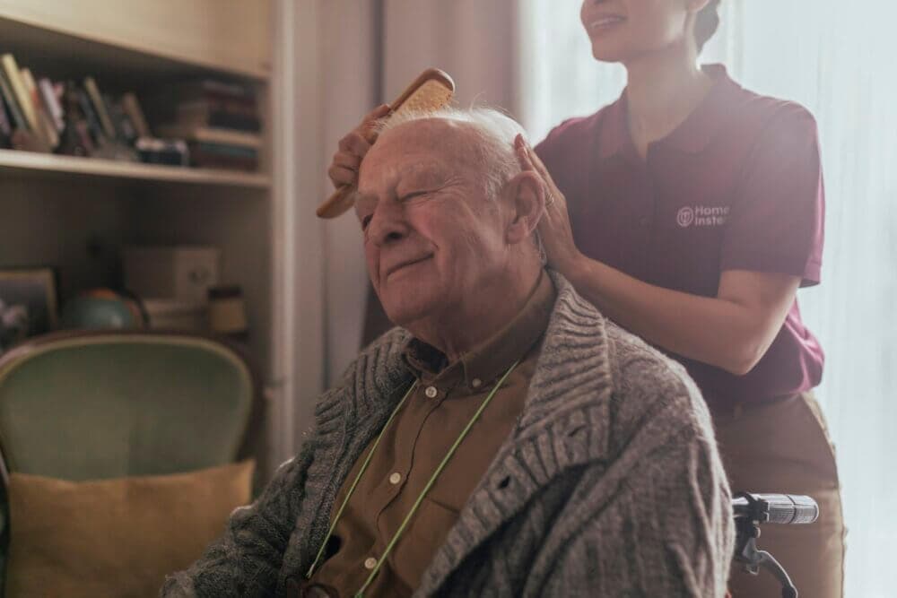 Elderly man in a wheelchair smiles while caregiver brushes his hair, a bookcase and chair in the background. - Home Instead