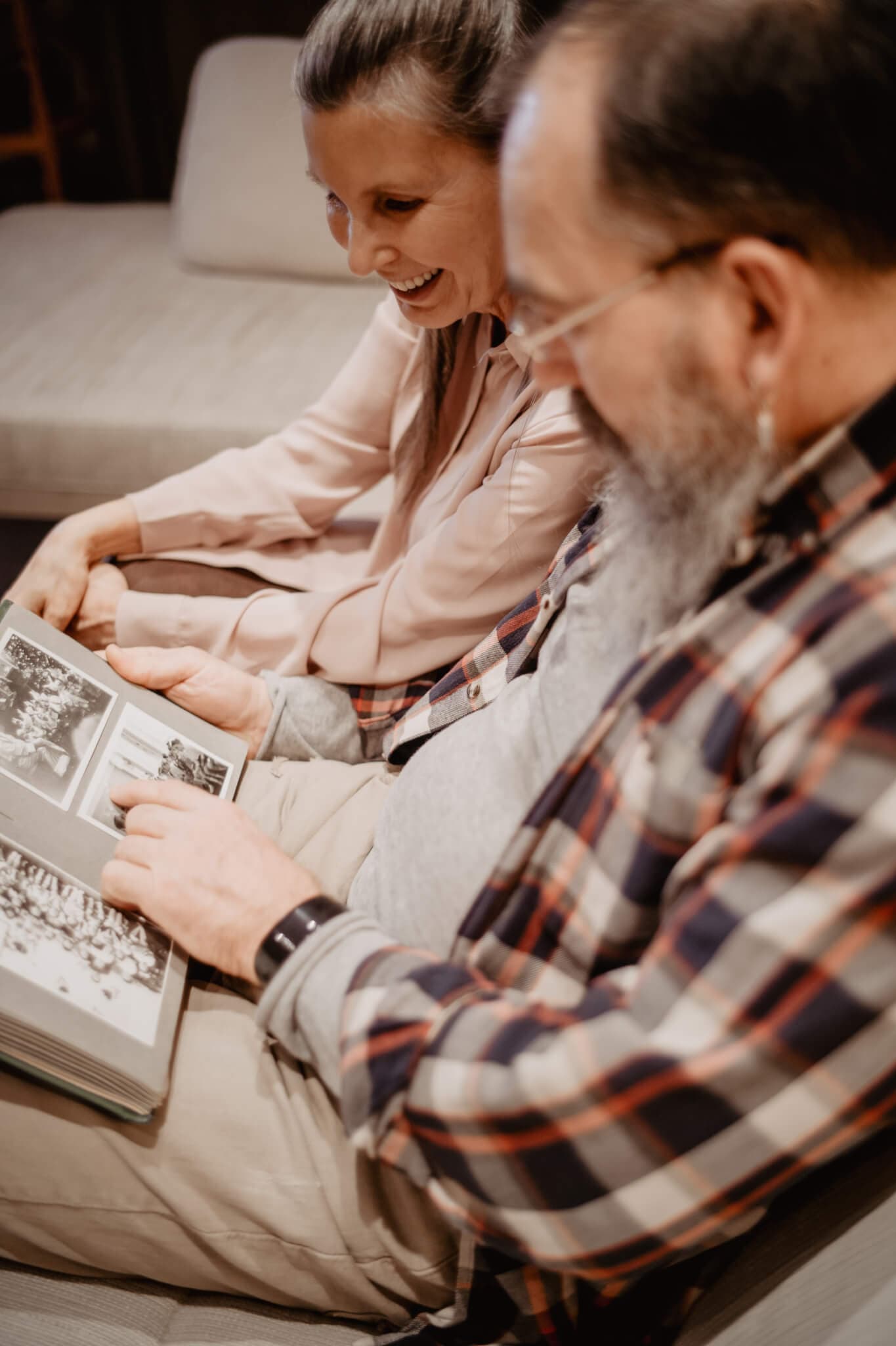 A smiling couple sits on a couch, looking through a photo album together. - Home Instead