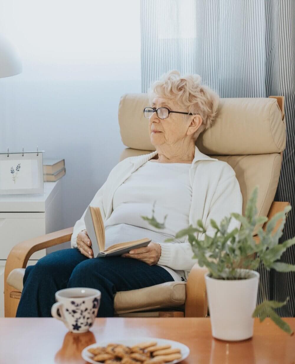Elderly person with glasses reading a book in an armchair, with a cup of tea and cookies on the table. - Home Instead