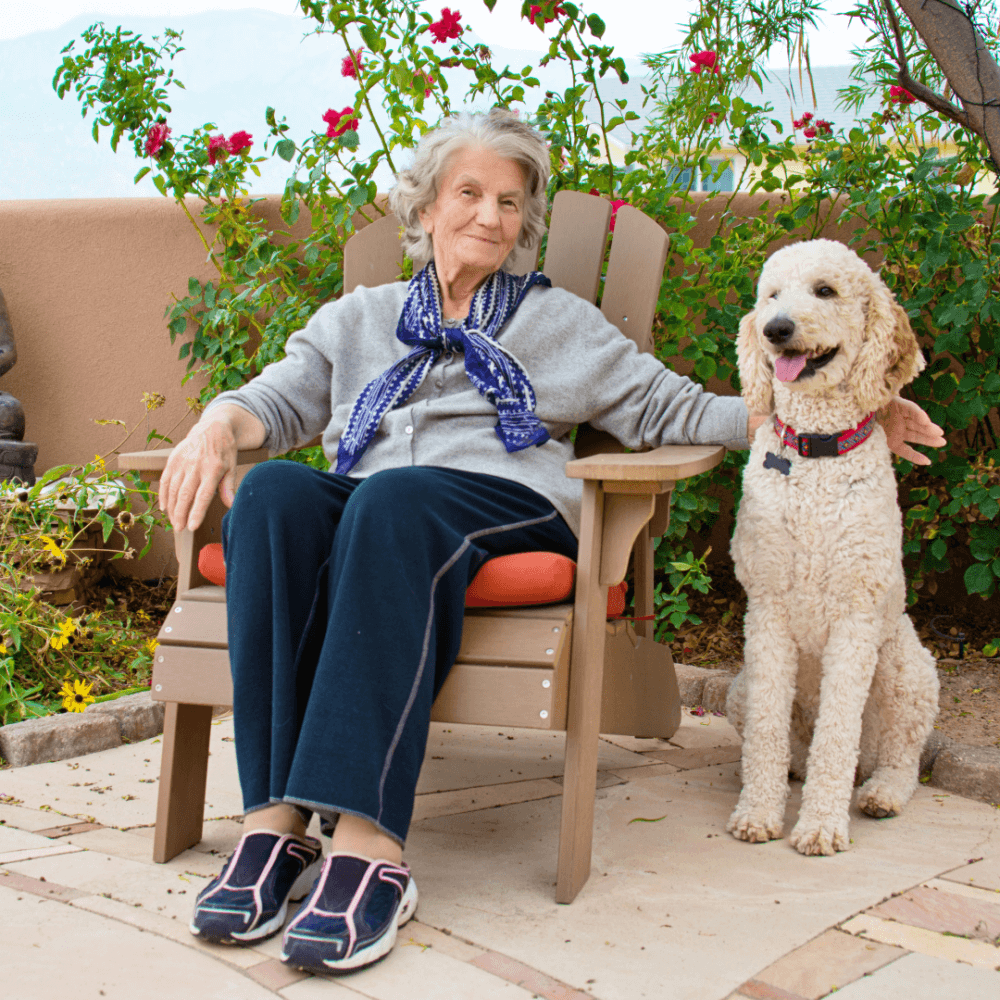 Elderly woman sitting in a chair outdoors, smiling with her arm around a fluffy dog beside her. - Home Instead