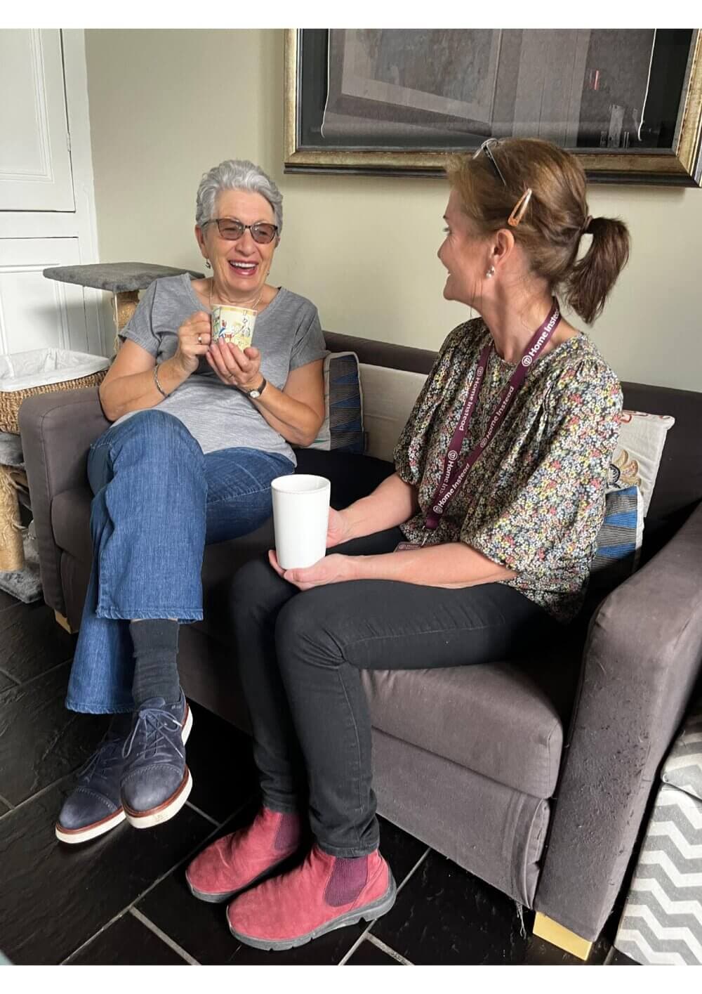 Two women sitting on a couch, smiling and holding mugs, engaged in a friendly conversation in a cozy room. - Home Instead
