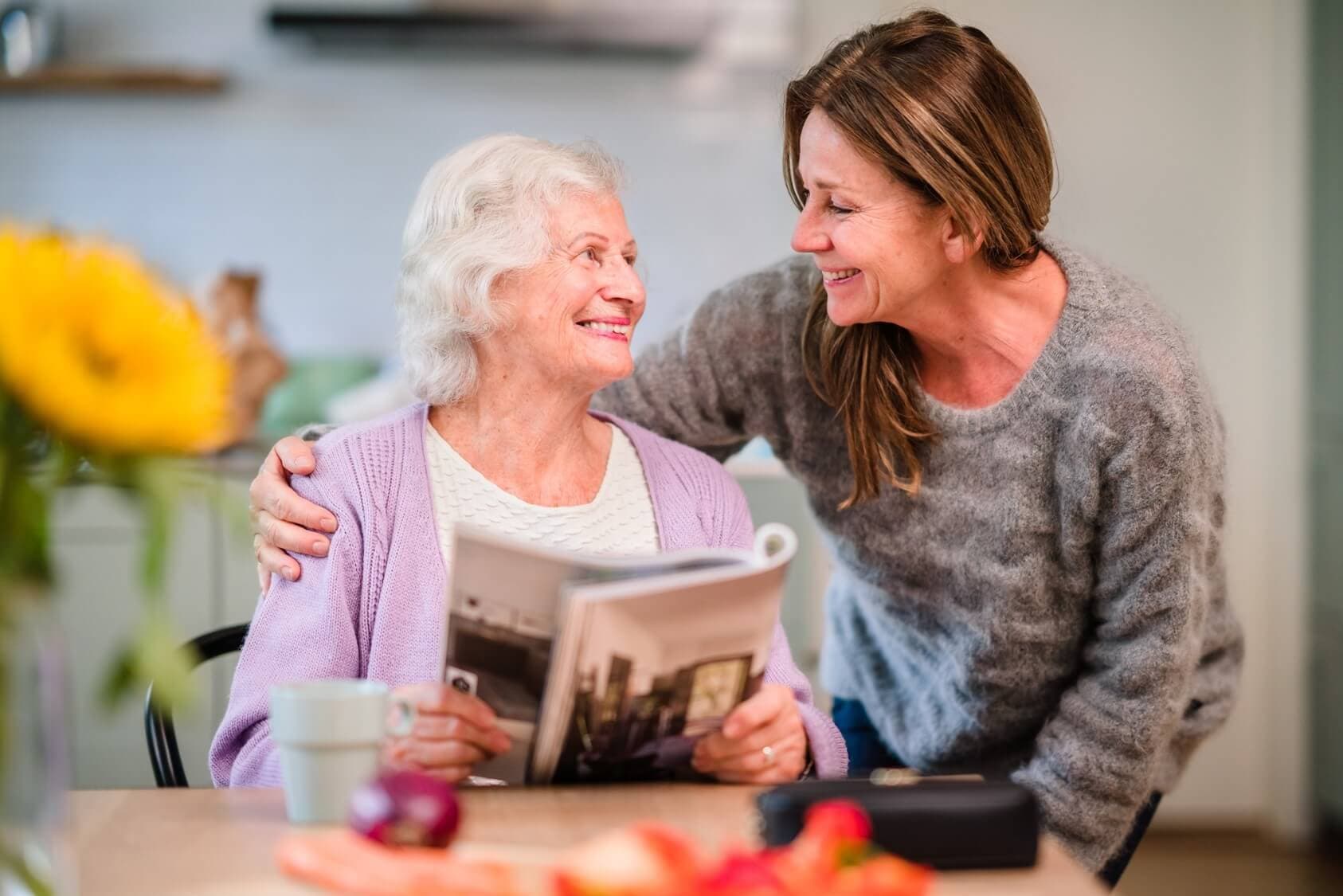 An elderly woman with a magazine smiles at a younger woman who has her arm around her in a cozy kitchen setting. - Home Instead