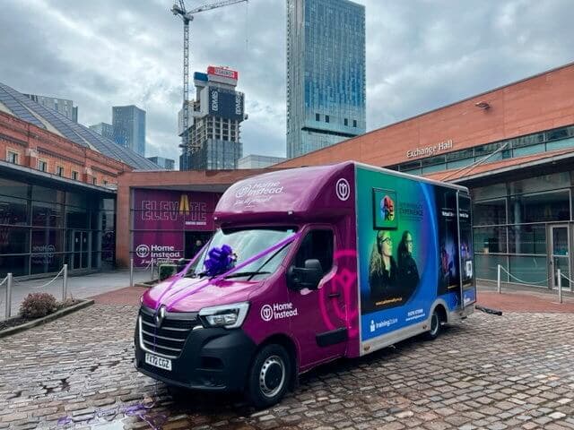A vibrant van with colorful graphics is parked on a cobblestone street near modern buildings and a construction crane. - Home Instead