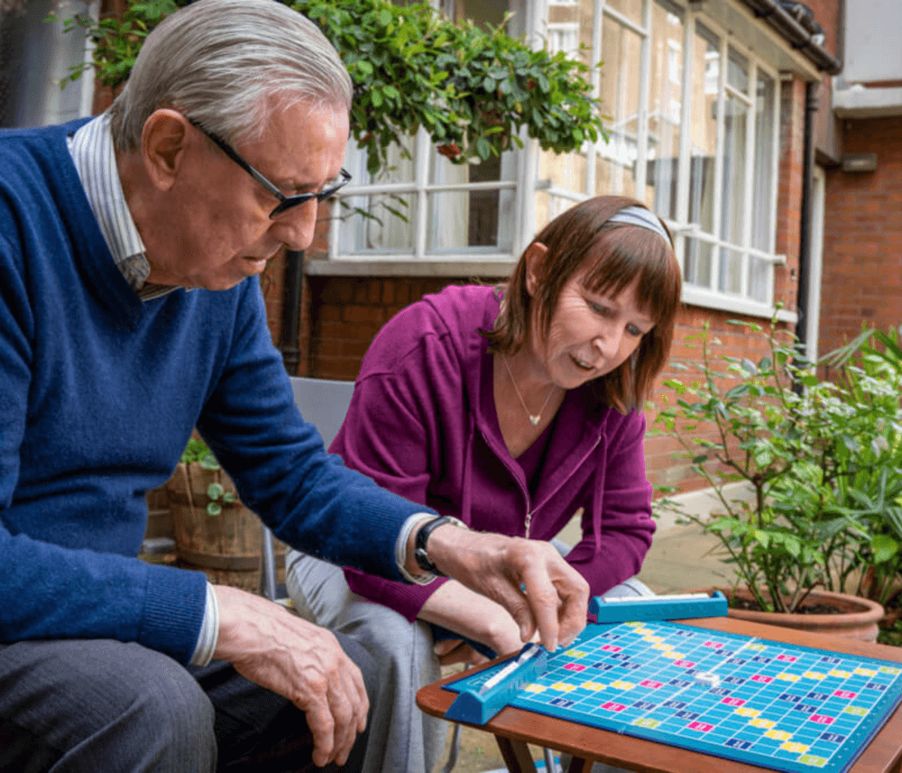 Two people are playing Scrabble together on an outdoor patio with plants and buildings in the background. - Home Instead