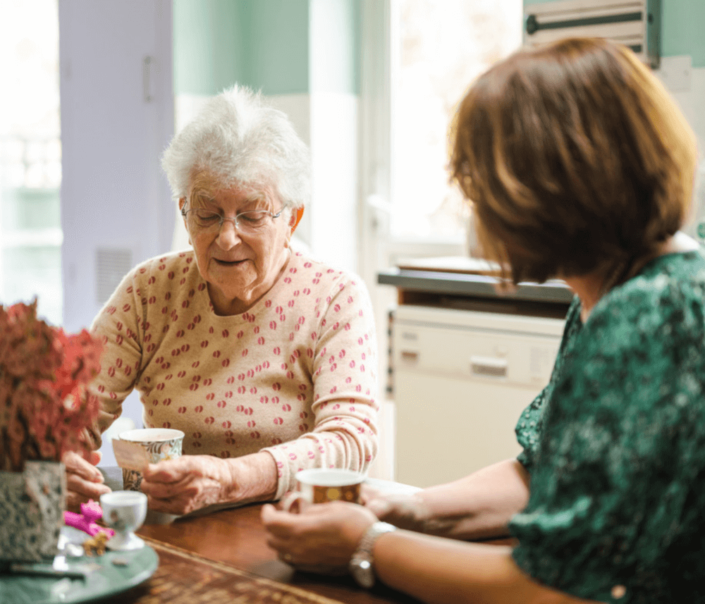 Elderly woman and younger woman sitting at a table, drinking tea and chatting in a cozy kitchen. - Home Instead