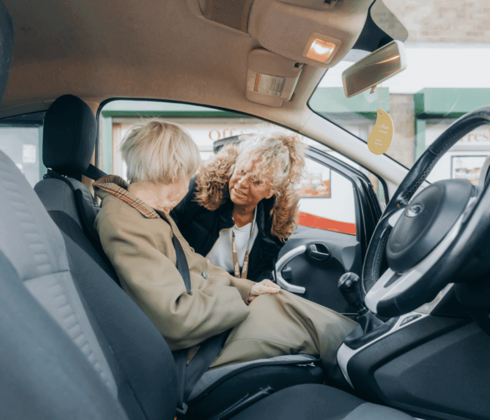 A woman helps an older adult seated in a car, possibly offering assistance or engaging in conversation. - Home Instead