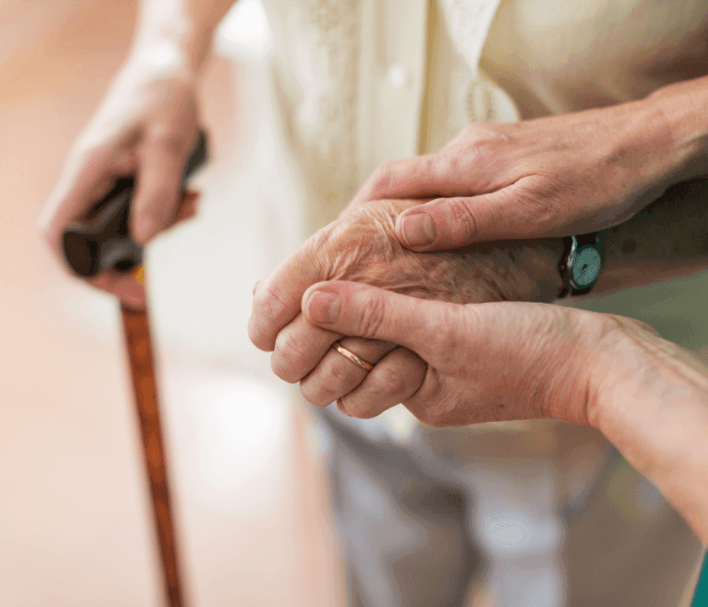 A close-up of a person holding the hand of an elderly individual who is using a wooden cane for support. - Home Instead