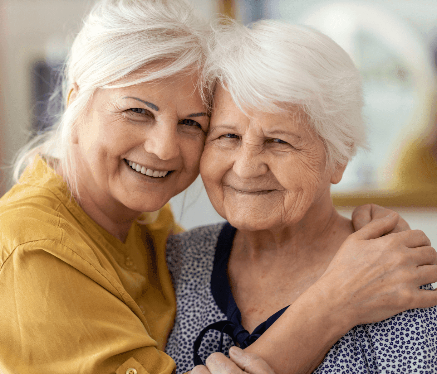 Two older women with white hair smiling and hugging indoors, one wearing a yellow top and the other a patterned blouse. - Home Instead