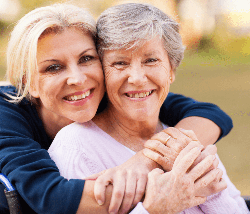 Two smiling women, one elderly and one middle-aged, embrace warmly outdoors. - Home Instead