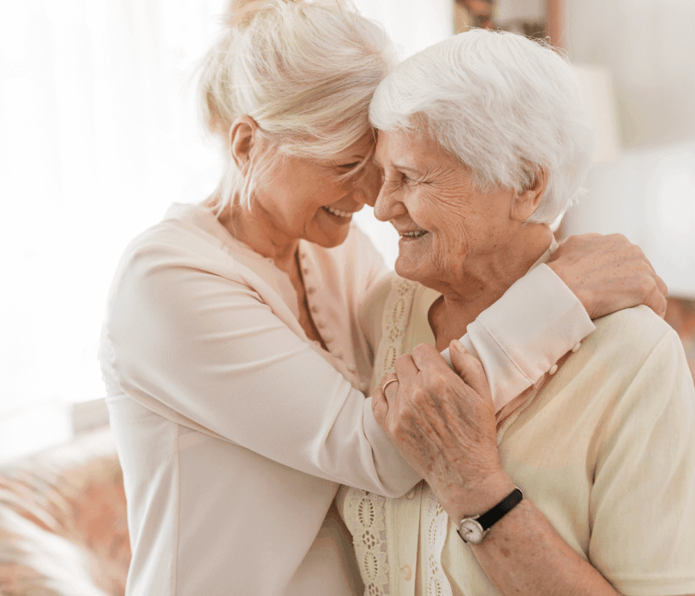 Two elderly women hug and smile at each other affectionately in a cozy living room. - Home Instead