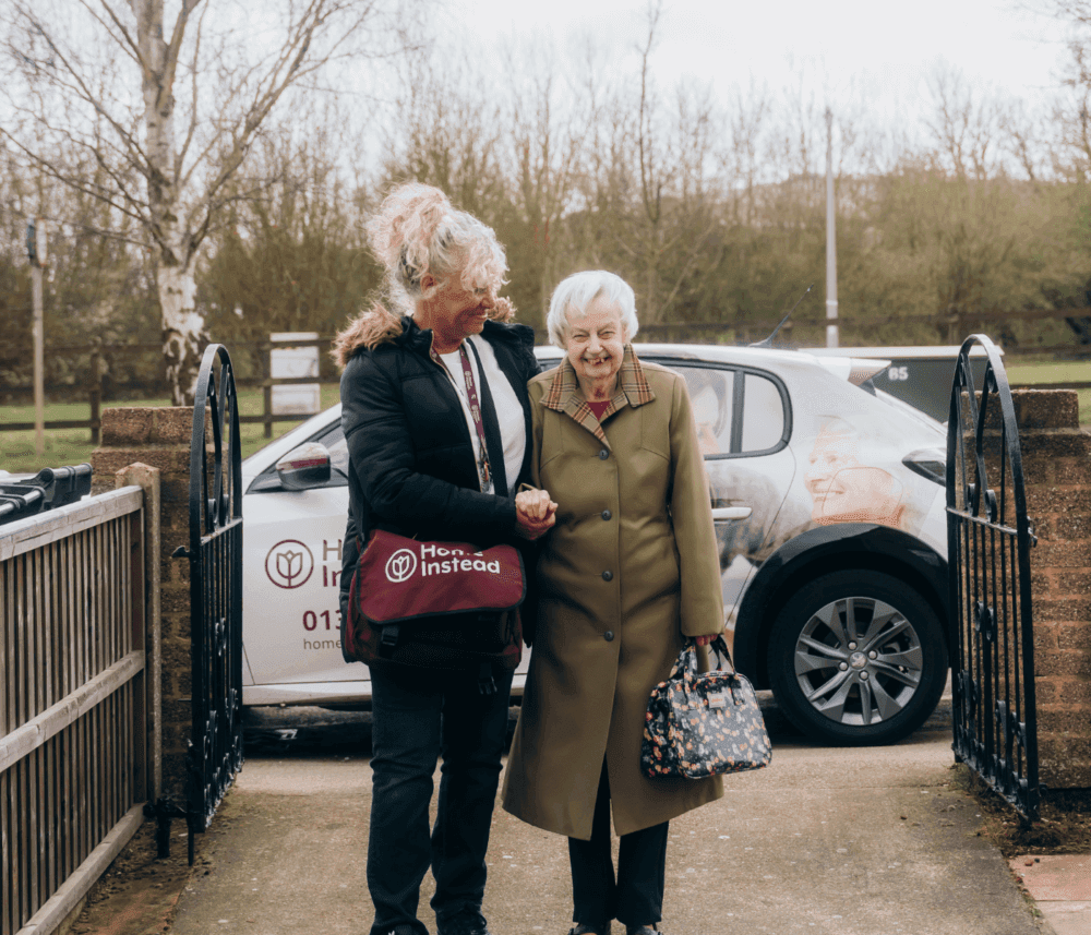 A caregiver and elderly woman smiling and walking arm in arm outside near a car parked in the background. - Home Instead