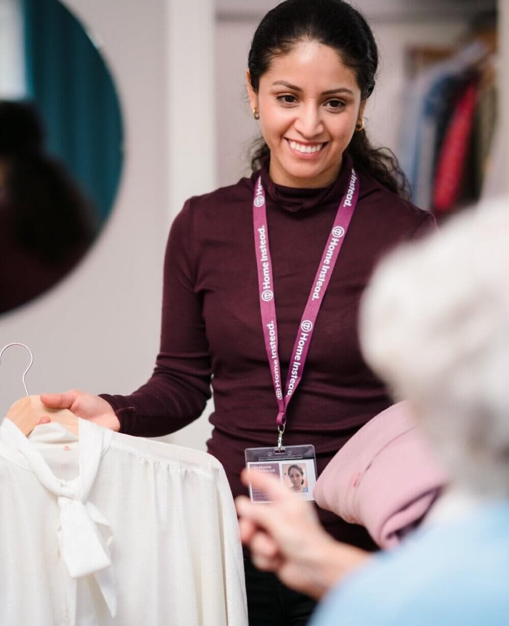 A smiling woman holds a white blouse on a hanger while talking to another person in a room with clothes. - Home Instead