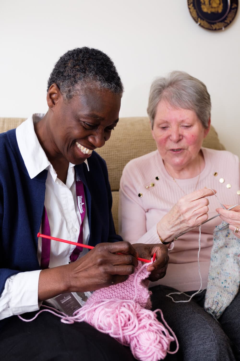 Client and Care Professional sit on a couch, smiling and knitting together with pink yarn. - Home Instead Bournemouth & Christchurch