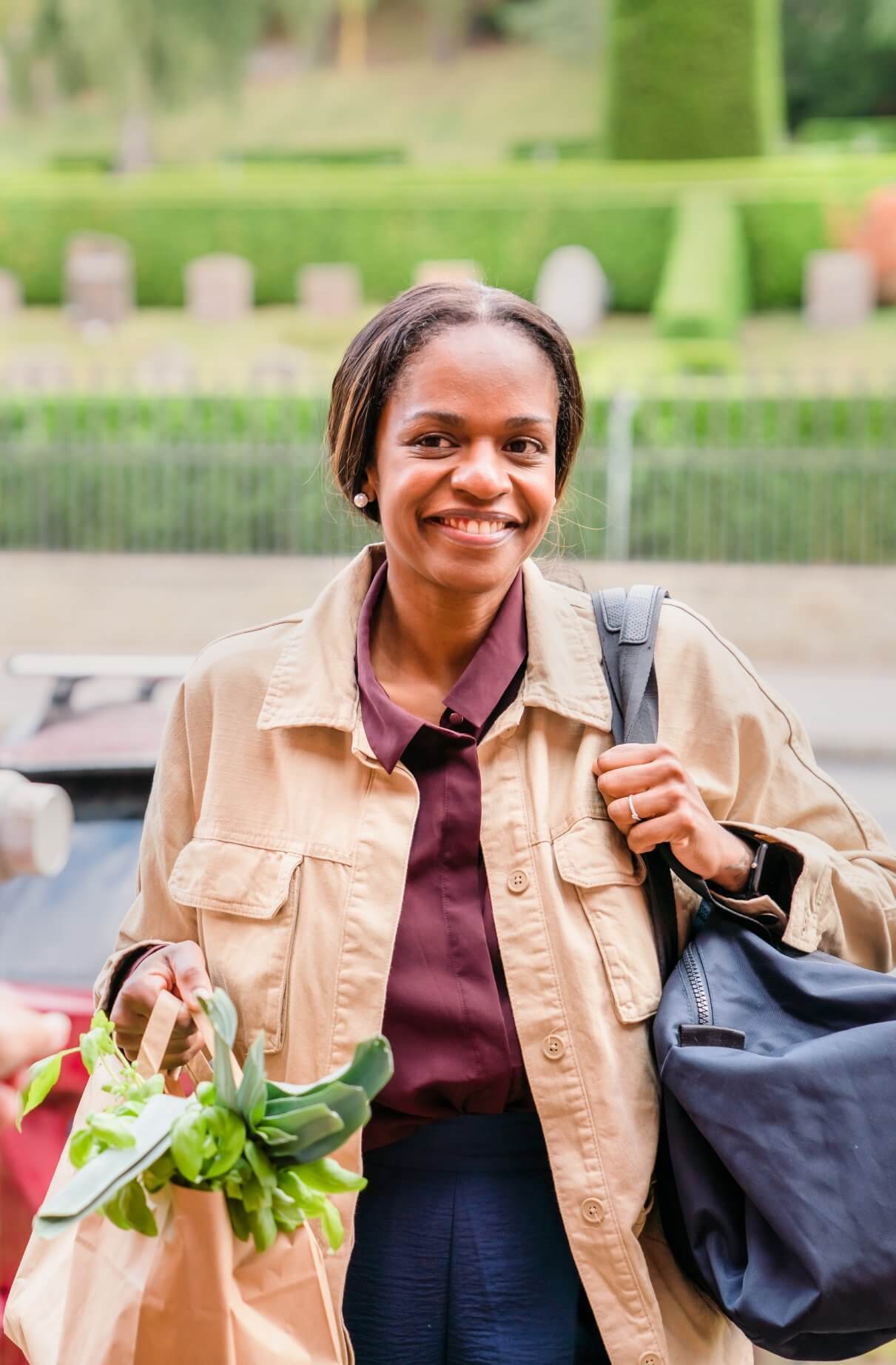 A woman smiles while holding grocery bags and a shoulder bag outdoors with greenery in the background. - Home Instead