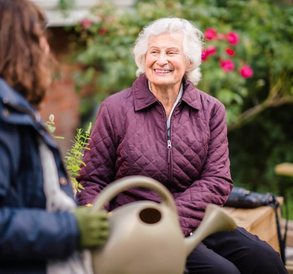 An elderly woman with white hair smiles while sitting outdoors, a person holding a watering can is partially visible. - Home Instead