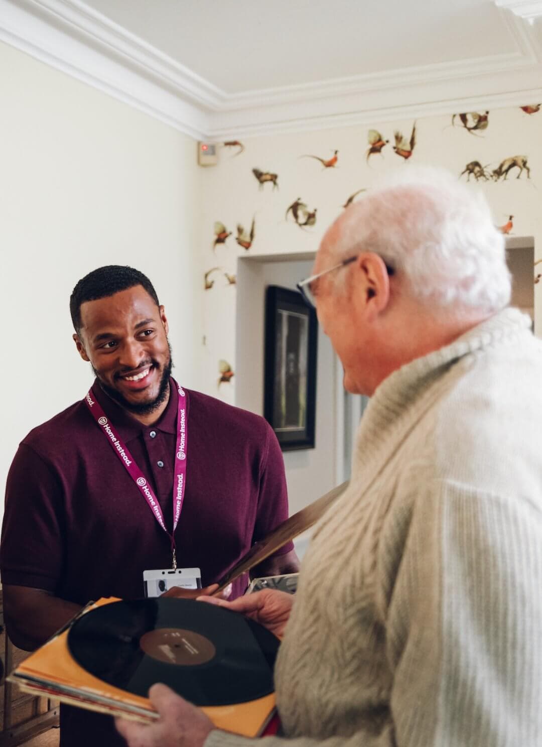 A younger man in a maroon shirt smiles as an older man in a beige sweater shows him a vinyl record. - Home Instead
